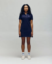 A woman with curly hair models the Psycho Bunny WOMENS POLO DRESS - WO2000003 in navy, styled with white sneakers and blue-striped socks, standing against a plain light gray background.