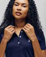 A woman with curly hair adjusts the collar of a navy blue Psycho Bunny WOMENS POLO DRESS - WO2000003, featuring an embroidered Bunny logo—a small skull and crossbones with bunny ears—on the chest.