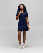 A person with long curly hair wears the Psycho Bunny WOMENS POLO DRESS - WO2000003, paired with white sneakers and striped socks, standing against a plain light gray background.