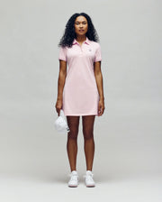 A person faces forward in a Psycho Bunny WOMENS POLO DRESS - WO2000003, a light pink polo dress with a feminine silhouette, styled with white sneakers and holding a white baseball cap against a plain light gray background.
