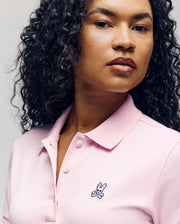 A woman with curly hair wears the Psycho Bunny WOMENS POLO DRESS - WO2000003 in light pink, featuring a feminine silhouette, Mother-of-Pearl buttons, and an embroidered bunny and crossbones logo on the chest.