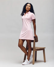 A woman with curly hair poses by a wooden chair, wearing the Psycho Bunny WOMENS POLO DRESS - WO2000003 in light pink with Mother-of-Pearl buttons and white sneakers, set against a plain light background.