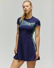 A woman models the Psycho Bunny TENNIS BALL CREW WOMENS DRESS ENGLISH (WO2000004), a short-sleeved navy tennis dress with light blue and green stripes plus a racket and ball graphic, standing against a light gray background.