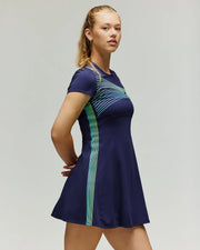 A woman stands confidently in the Psycho Bunny TENNIS BALL CREW WOMENS DRESS ENGLISH - WO2000004, a navy short-sleeve tennis dress with blue and green diagonal stripes, against a plain light background.