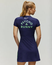A woman with long blonde hair in a ponytail wears the Psycho Bunny Tennis Ball Crew Womens Dress WO2000004, a fitted navy blue piece featuring “PSYCHO BUNNY” and a small green bunny graphic on the back, standing with her back to the camera.