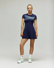A woman faces forward with a neutral expression, wearing the Psycho Bunny TENNIS BALL CREW WOMENS DRESS ENGLISH - WO2000004 in navy blue with light blue and green accents, paired with white socks and sneakers against a plain backdrop. One leg is slightly bent.