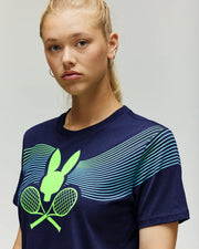 A young woman with blonde hair in a ponytail wears the Psycho Bunny TENNIS BALL CREW WOMENS TEE, featuring a neon green bunny head, crossed tennis rackets, and curved blue and green lines across the chest.