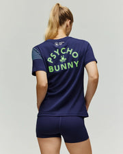 A woman with a ponytail faces away, wearing the Psycho Bunny Tennis Ball Crew Womens Tee, featuring "PSYCHO BUNNY" in neon green on the back, light blue stripes on one sleeve, and paired with matching navy shorts.