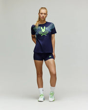 A woman stands against a plain background wearing the Psycho Bunny Tennis Ball Crew Womens Tee in navy (WT0200003), matching navy shorts, white socks, and white sneakers.