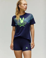 A woman stands against a plain light background, looking to the side, in the Psycho Bunny TENNIS BALL CREW WOMENS TEE FRENCH (WT0200004), a navy blue Pima cotton tee with neon green Tennis Bunny graphics, paired with matching shorts.