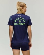 A woman with blonde hair in a ponytail is seen from behind wearing the Psycho Bunny TENNIS BALL CREW WOMENS TEE FRENCH (WT0200004), a navy Pima cotton tee with a neon green Tennis Bunny graphic on the back, paired with navy shorts.
