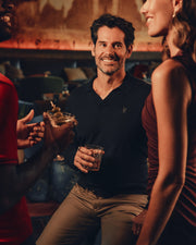 A man in a Psycho Bunny MENS JOHNNY COLLAR SWEATER - B6E258E200 and beige pants holds a drink and smiles, surrounded by two people chatting in a warmly lit bar or lounge.