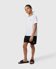 A young person stands against a plain white background, wearing a casual white t-shirt, KIDS MALTA HYDROCHROMIC SWIM TRUNK - B0W321B2SW from Psycho Bunny, and black slide sandals. Their hair is styled in cornrows, and they have a confident expression on their face.