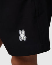 Close-up of the hem of a pair of Psycho Bunny KIDS MALTA HYDROCHROMIC SWIM TRUNK - B0W321B2SW, featuring a small bunny head logo with crossed bones underneath. The logo is white with a gray outline and stands out against the black fabric. The subtle bunny pattern offers a unique touch, while the background remains plain and unobtrusive.