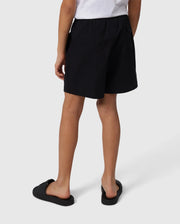 A person wearing a white T-shirt with a subtle bunny pattern stands facing away, showcasing the KIDS MALTA HYDROCHROMIC SWIM TRUNK in black by Psycho Bunny and black slip-on shoes against a neutral background. Their hands rest loosely at their sides.