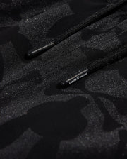 Close-up of Psycho Bunny's KIDS MALTA HYDROCHROMIC SWIM TRUNK - B0W321B2SW, showcasing a black textured fabric with a subtle camouflage pattern in shades of dark grey and black. Two parallel black cords with small text on one cord are partially visible, reminiscent of sleek swim trunks. The overall appearance is both modern and hydrochromic.