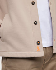 Close-up of a person sporting the elevated Psycho Bunny beige MENS BEDFORD SHIRT JACKET - B6J290B200 with black buttons, layered over a white shirt and tan pants. The jacket features an orange embroidered bunny logo near the bottom. One hand is partially visible inside the jacket's pocket, highlighting its exceptional on-body feel.
