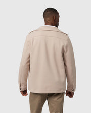 A man is standing with his back to the camera, wearing a stylish beige MENS BEDFORD SHIRT JACKET - B6J290B200 by Psycho Bunny, featuring a collar, button cuffs, and shoulder straps. He has paired it with khaki pants against a plain, light gray background.