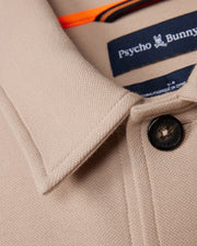 Close-up of the Psycho Bunny MENS BEDFORD SHIRT JACKET - B6J290B200, showcasing its beige fabric with a distinctive black button and the iconic "Psycho Bunny" logo label. The textured material emphasizes its high-quality craftsmanship, while contrasting elements add a stylish touch. An orange stripe inside the collar mimics work-jacket pocket flair for added character.