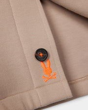 Close-up of a beige fabric with a black button stitched onto it. Below the button, there is an orange embroidered logo featuring Psycho Bunny's iconic bunny head with crossed bones underneath. The edge of the fabric is neatly hemmed, showcasing the exceptional on-body feel and elevated style of the MENS BEDFORD SHIRT JACKET - B6J290B200 by Psycho Bunny.