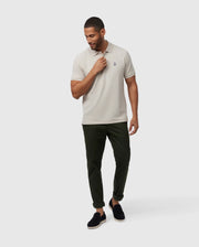 A man in a Psycho Bunny MENS CLASSIC PIQUE POLO SHIRT - B6K001B200, light beige, paired with dark green pants stands against a white background. He wears black slip-on shoes and has neat short hair. His left hand touches his neck while he gazes downwards, showcasing men's fashion.