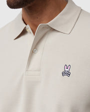 A close-up of someone wearing the Psycho Bunny MENS CLASSIC PIQUE POLO SHIRT - B6K001B200 in light beige, crafted from Pima cotton with two buttons undone. It features a small pink and white bunny with crossbones logo on the chest, representing timeless men's fashion.