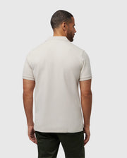 A man faces away from the camera, wearing a Psycho Bunny MENS CLASSIC PIQUE POLO SHIRT - B6K001B200 in light gray with dark pants. The simple white backdrop and his subtly turned head capture the essence of timeless men's fashion.