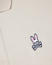 A close-up of the Psycho Bunny beige MENS CLASSIC PIQUE POLO SHIRT - B6K001B200, made from premium Pima cotton, showcases a small bunny skull emblem with pink ears and crossbones. It adds an edgy twist to men's fashion.