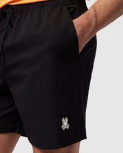 A person wearing the MENS MALTA HYDROCHROMIC SWIM TRUNK by Psycho Bunny, featuring a small, stylized bunny logo on the front, places their hand in their pocket. The background is plain white, and the person's upper body and face are not shown.