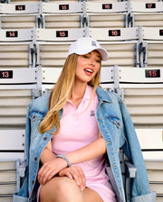A woman with long blonde hair sits in empty stadium seats, smiling and posing for the camera in a Psycho Bunny WOMENS POLO DRESS (WO2000003), white cap, and blue denim jacket draped over her shoulders.