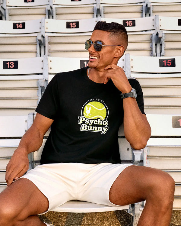 A man in sunglasses and a Psycho Bunny MENS PETE TENNIS GRAPHIC TEE (MT0200012) paired with white shorts sits smiling on stadium seats, his left arm on the backrest and right hand touching his chin.