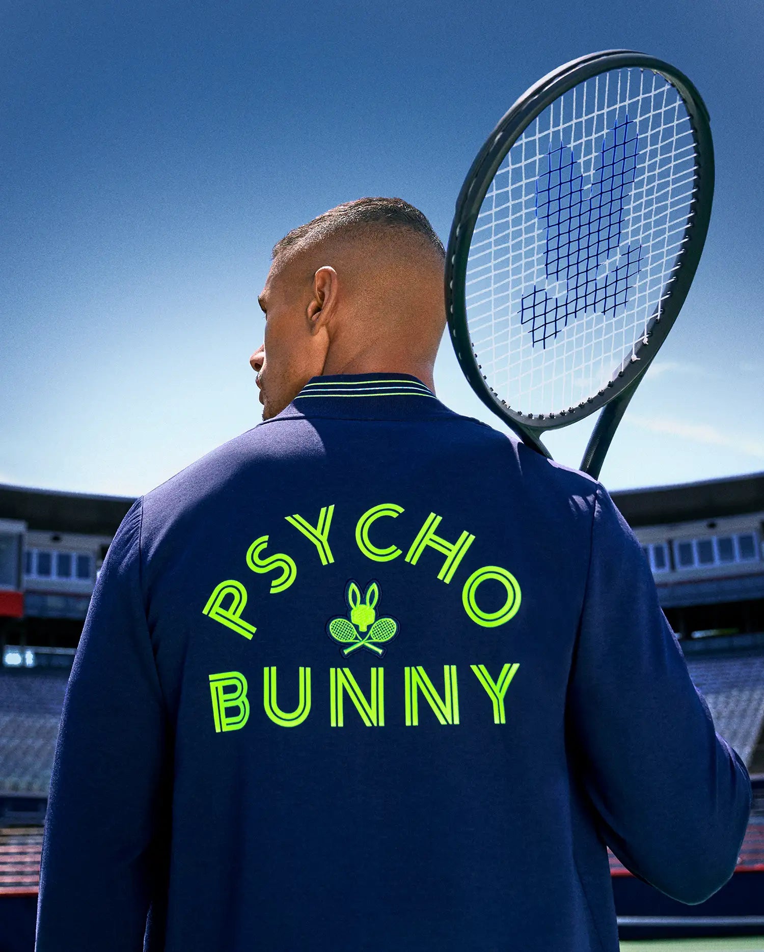 On a tennis court, a man holds his racket over his shoulder, showing the back of his Psycho Bunny TENNIS BALL CREW MENS JACKET ENGLISH, detailed with neon yellow "PSYCHO BUNNY" text and an embroidered Bunny logo with crossed rackets.