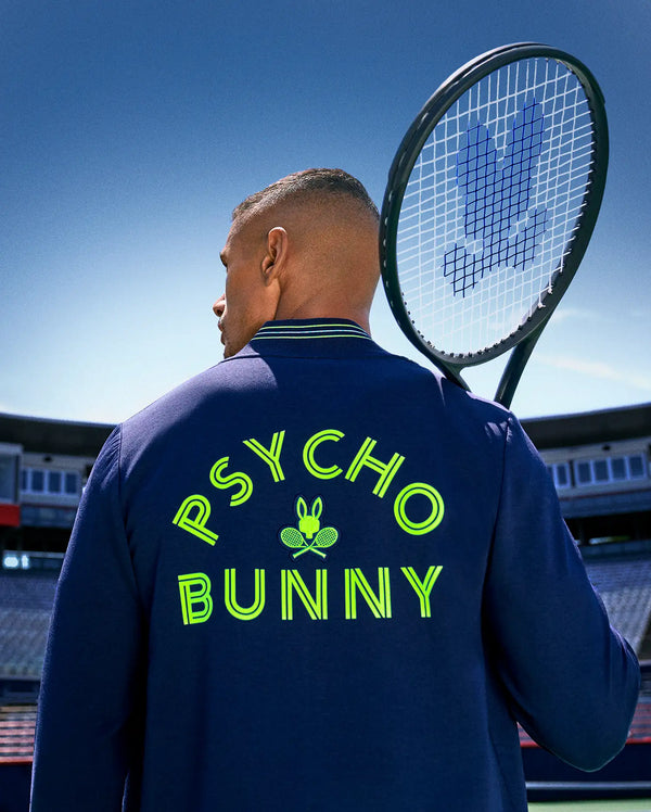 On a tennis court, a man holds his racket over his shoulder, showing the back of his Psycho Bunny TENNIS BALL CREW MENS JACKET ENGLISH, detailed with neon yellow "PSYCHO BUNNY" text and an embroidered Bunny logo with crossed rackets.