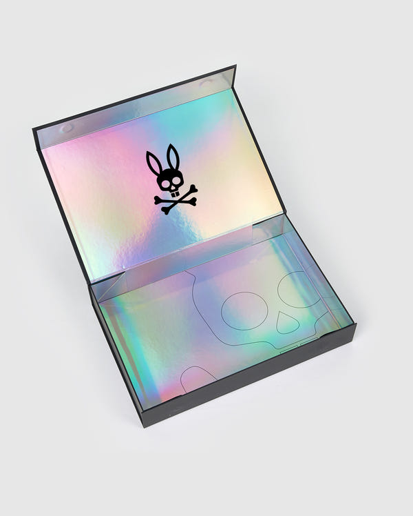 A sleek, iridescent GIFT BOX - GIFTBOX by Psycho Bunny is open, revealing a holographic interior. The lid features a black rabbit head logo with crossed bones beneath it. The base of the box has faint outlines of a skull design, and the exterior is black with magnetic closures.
