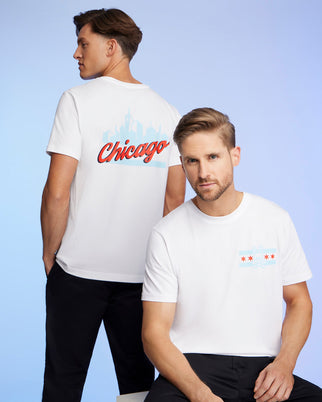 Two men wear Chicago-themed Psycho Bunny tees made from soft Pima cotton. One displays "Chicago" and the skyline on his back, while the other features the city's flag and skyline on the front. The signature Bunny logo adorns each shirt against a calming blue background.