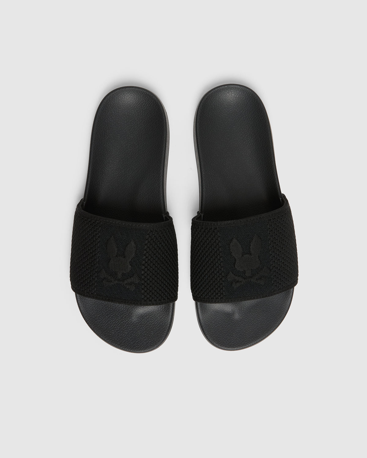 A pair of black Psycho Bunny KIDS KNIT SLIDES - B0A377F200, featuring a textured strap with an embroidered stylized rabbit head logo and a molded footbed for comfort, are placed side by side on a light gray background.