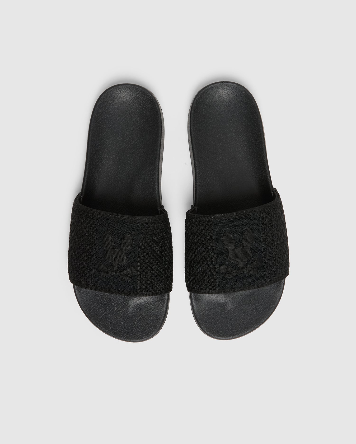 A pair of black Psycho Bunny KIDS KNIT SLIDES - B0A377F200, featuring a textured strap with an embroidered stylized rabbit head logo and a molded footbed for comfort, are placed side by side on a light gray background.