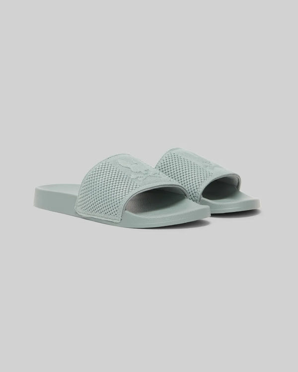 The Psycho Bunny KIDS KNIT SLIDES - B0A377F200 are light blue-green knit pool slides with textured open-toe straps, flat soles, and an ergonomically molded footbed, shown against a plain light gray background.