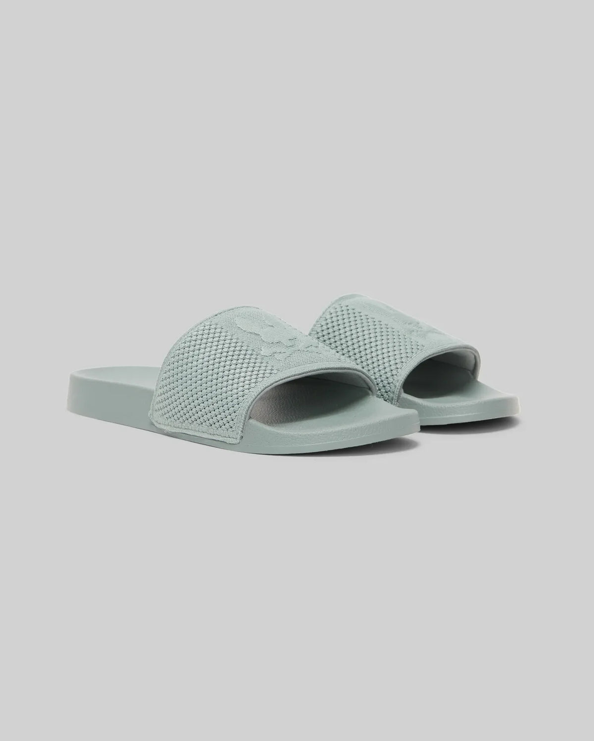 The Psycho Bunny KIDS KNIT SLIDES - B0A377F200 are light blue-green knit pool slides with textured open-toe straps, flat soles, and an ergonomically molded footbed, shown against a plain light gray background.