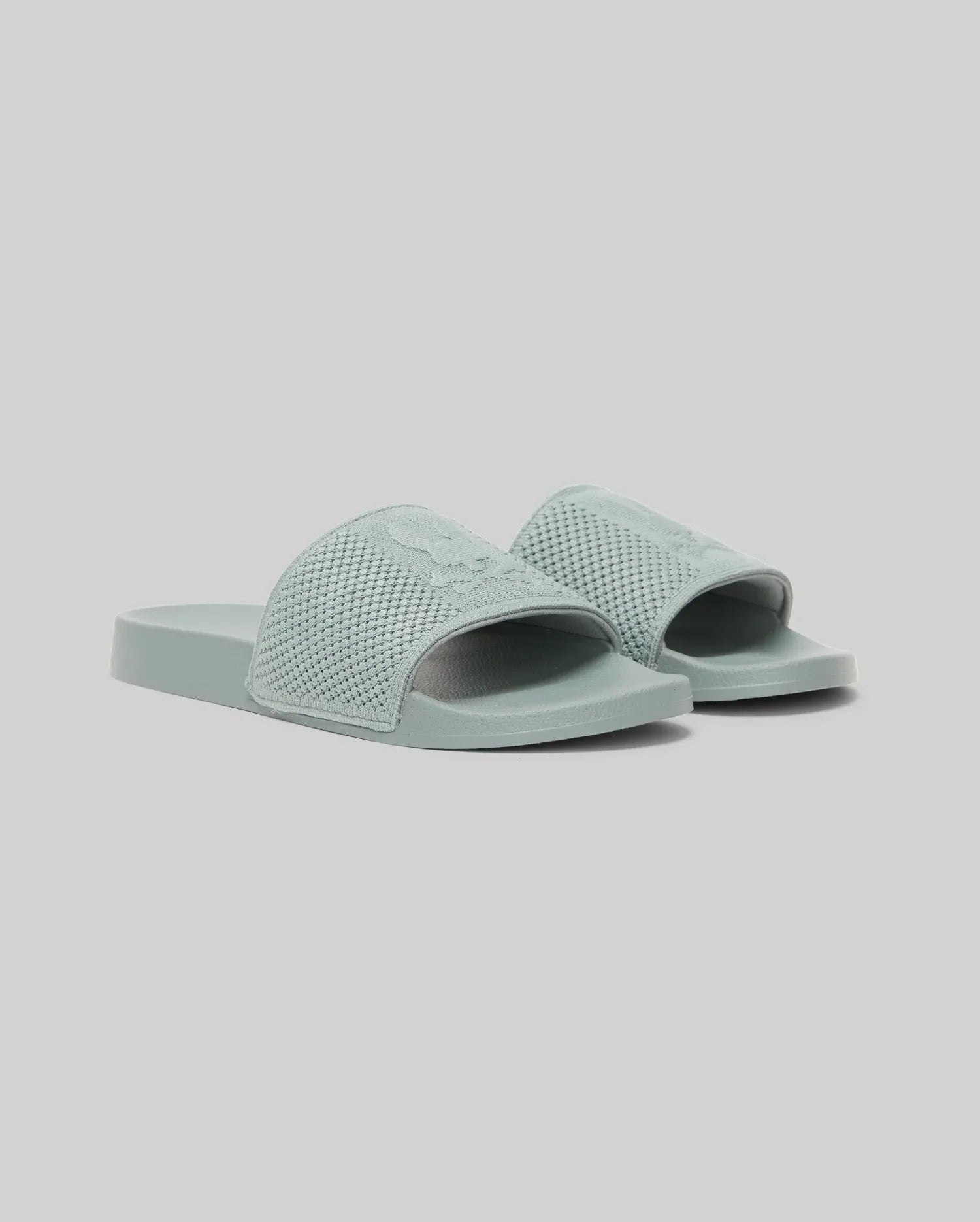 The Psycho Bunny KIDS KNIT SLIDES - B0A377F200 are light blue-green knit pool slides with textured open-toe straps, flat soles, and an ergonomically molded footbed, shown against a plain light gray background.