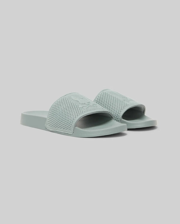 The Psycho Bunny KIDS KNIT SLIDES - B0A377F200 are light blue-green knit pool slides with textured open-toe straps, flat soles, and an ergonomically molded footbed, shown against a plain light gray background.