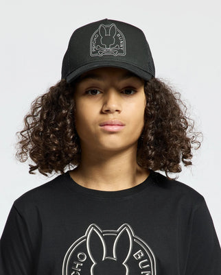 A person with curly hair dons a black KIDS MELBOURNE GRAPHIC TRUCKER HAT (B0A548F200) by Psycho Bunny, featuring an embroidered bunny logo, paired with a black shirt also showcasing the brand's iconic emblem. They stand against a plain white background.