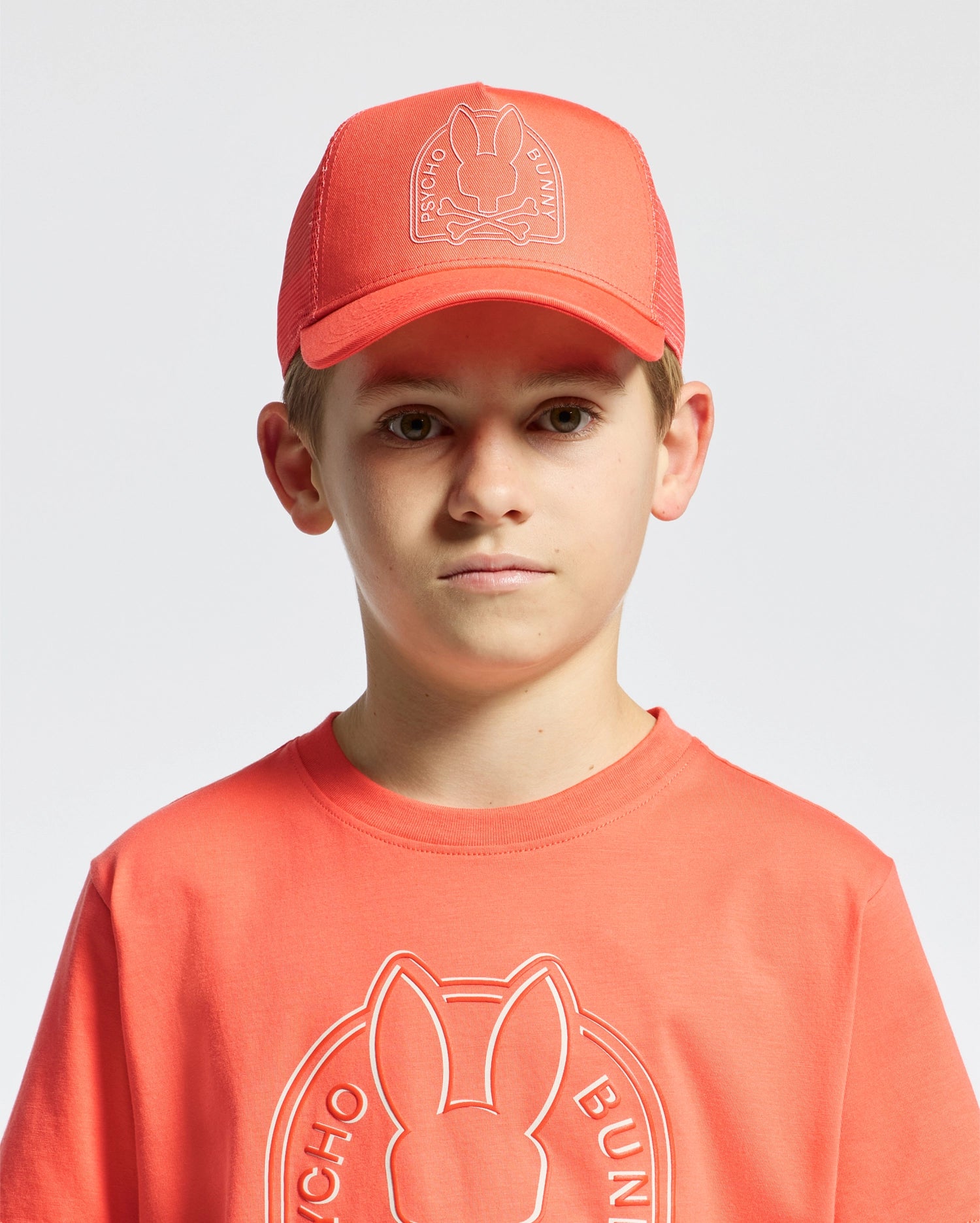 A young person sports a Psycho Bunny KIDS MELBOURNE GRAPHIC TRUCKER HAT - B0A548F200 and a matching t-shirt featuring an embroidered rabbit icon. The background is plain white.