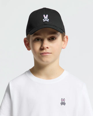 A young boy wears the Psycho Bunny KIDS CLASSIC BASEBALL CAP - B0A816A1HT, featuring a black design with an embroidered Bunny and crossbones logo. His plain white t-shirt complements the classic look against a simple gray background.