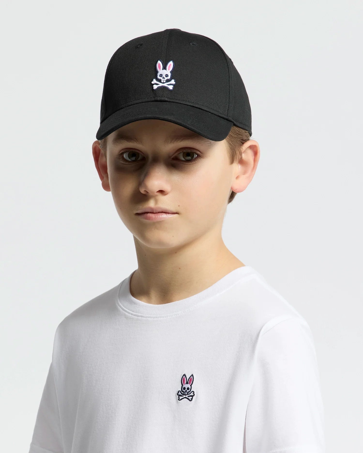 A young person wears a Psycho Bunny black KIDS CLASSIC BASEBALL CAP (B0A816A1HT) with an embroidered bunny and crossbones, complemented by a white T-shirt featuring the same bold logo. The plain white background accentuates their edgy style as they look at the camera.