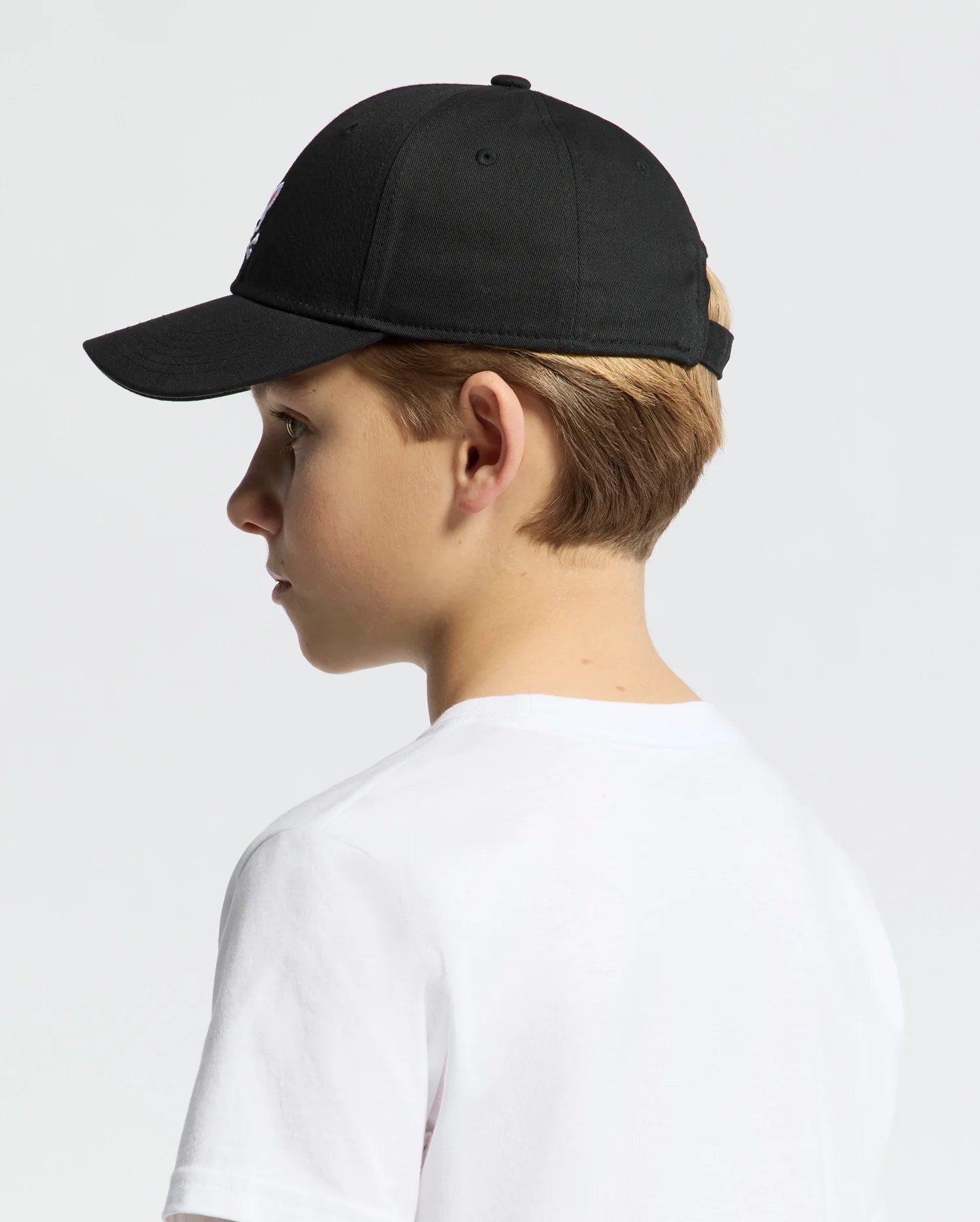 A youth wearing the Psycho Bunny KIDS CLASSIC BASEBALL CAP in black, product code B0A816A1HT, with an adjustable fit and an embroidered Bunny logo on the left side, paired with a classic white T-shirt against a simple light-colored background.