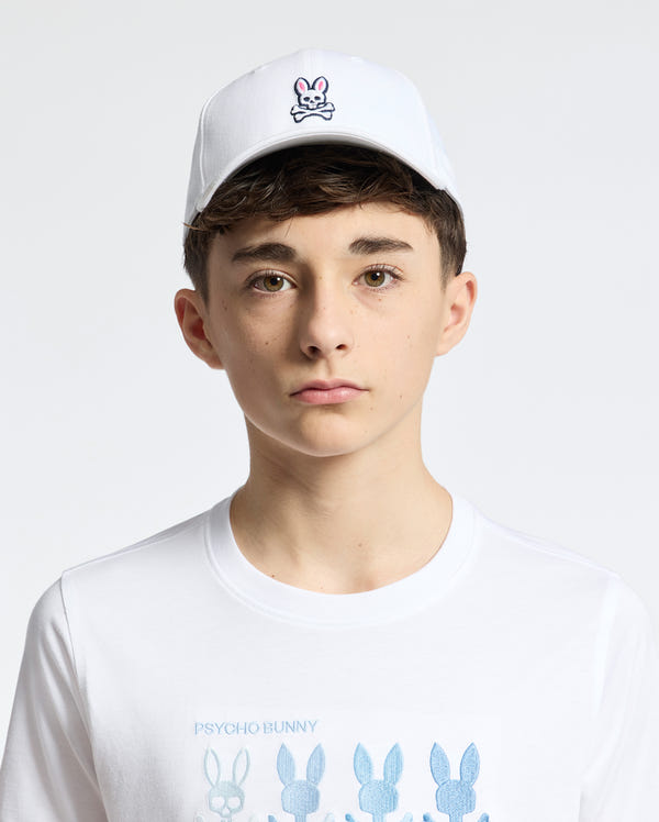 A teen boy wears a Psycho Bunny KIDS CLASSIC BASEBALL CAP (B0A816A1HT) in white with an embroidered pink and black Bunny logo, paired with a T-shirt featuring blue bunny silhouettes and "Psycho Bunny" text. The cap is adjustable for comfort against a plain white backdrop.