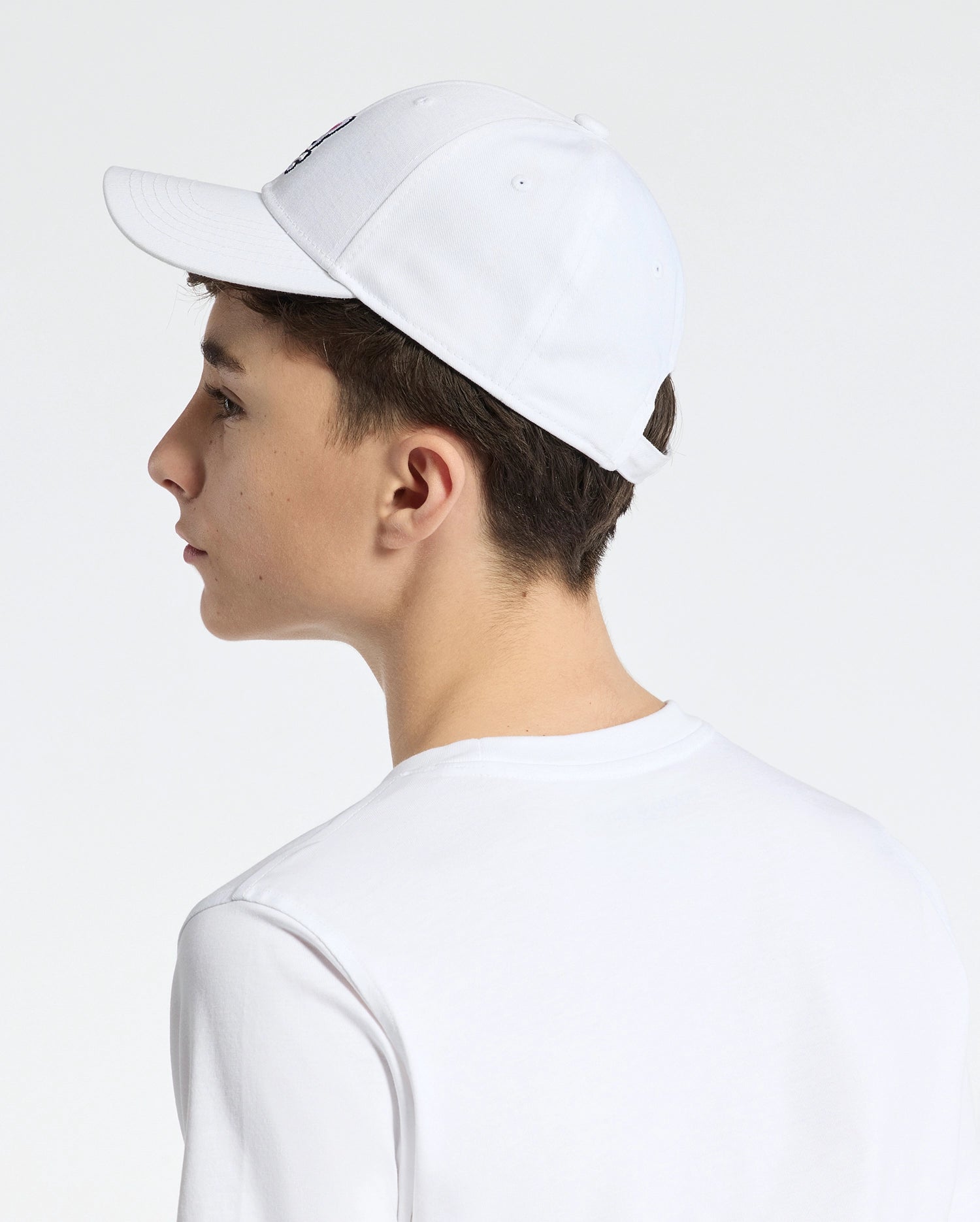 A youth in profile wears the Psycho Bunny KIDS CLASSIC BASEBALL CAP - B0A816A1HT, featuring an embroidered bunny. The white cap and matching T-shirt create a classic look as they gaze left against a neutral backdrop.