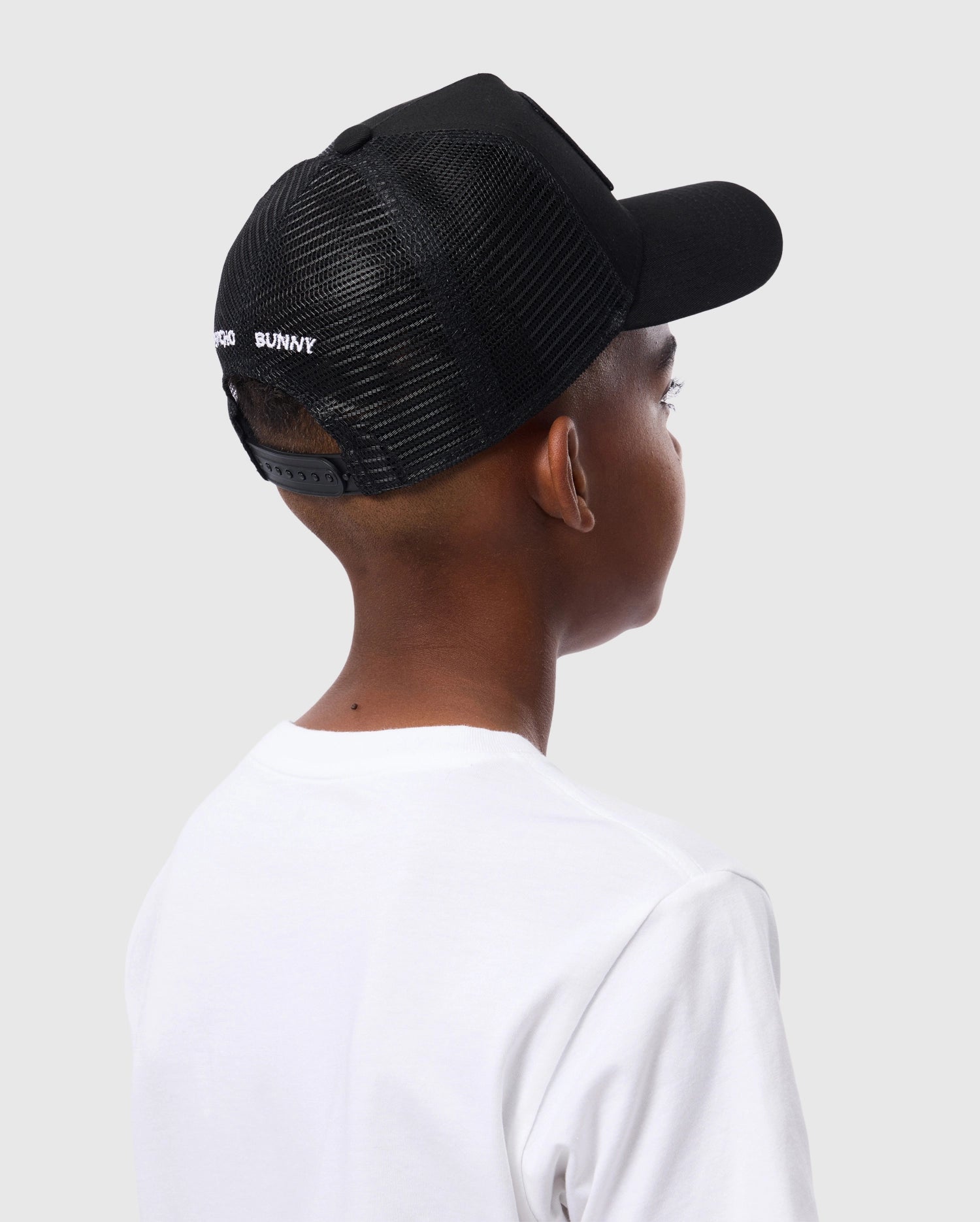 A child is seen from behind at an angle, wearing a Psycho Bunny KIDS BUNNY PATCH TRUCKER CAP (B0A965C200) with black breathable mesh paneling and a white t-shirt against a plain light gray background.