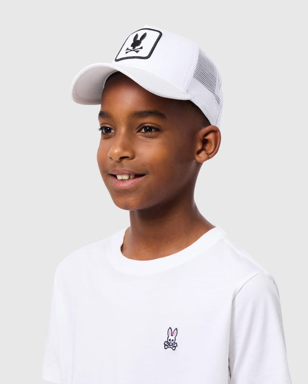 A boy in a Psycho Bunny KIDS BUNNY PATCH TRUCKER CAP - B0A965C200 poses against a plain background. The white cap with the playful Bunny logo patch pairs well with his white T-shirt, featuring a small embroidered cartoon character.